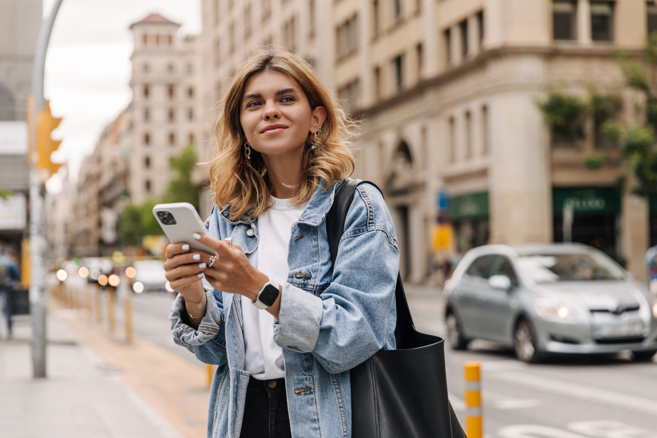 Picture of pretty young woman staying on the street holding phone in hands. Stylish blonde looking away and smiling in city.